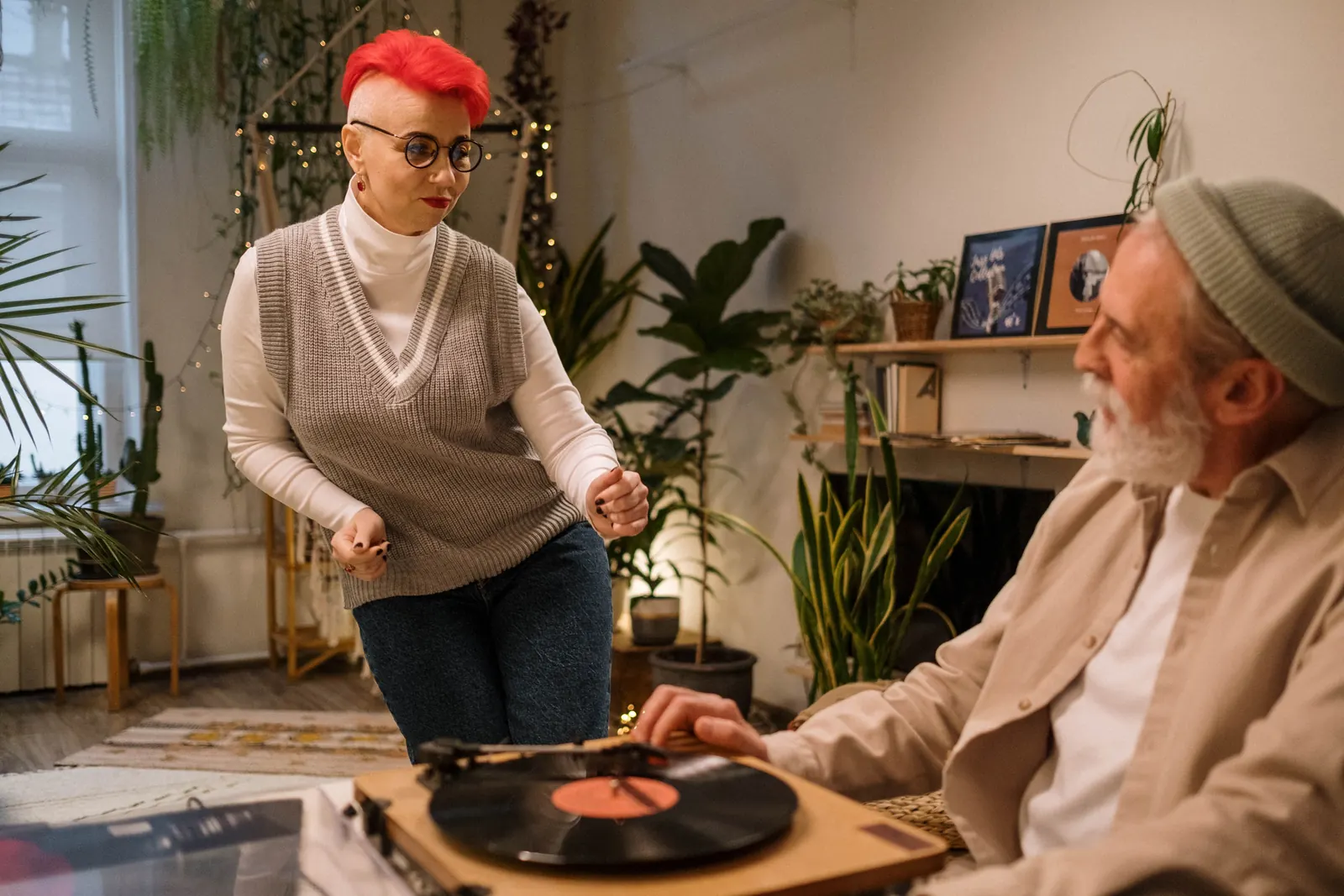 A woman with red hair dancing beside a record player in her first home, surrounded by plants